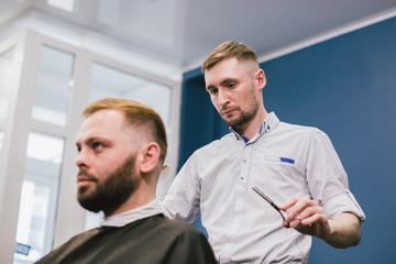Fototapeta na wymiar Barber Making Haircut Bearded Man In Barbershop. Professional stylist cutting client hair in salon. Barber using scissors and comb. Skillful hairdresser cutting male hair. Hair Care Service Concept