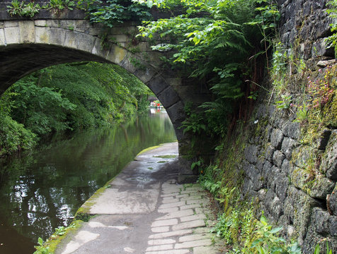 a path along the rochdale canal crossing under an old stone bridge overgrown with vegetation with trees and a narrowboat in the distance