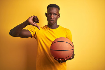 African american athlete man holding basketball ball standing over isolated yellow background with angry face, negative sign showing dislike with thumbs down, rejection concept