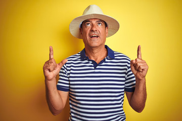 Handsome middle age man wearing striped polo and hat over isolated yellow background amazed and surprised looking up and pointing with fingers and raised arms.