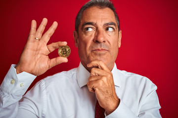Handsome middle age man holding bitcoin standing over isolated red background serious face thinking about question, very confused idea
