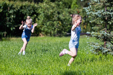 Naklejka na ściany i meble Children run on the green grass holding hands. Girls running around the lawn with grass playing with splashes of water to water plants.