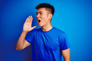 Young asian chinese man wearing t-shirt standing over isolated blue background shouting and screaming loud to side with hand on mouth. Communication concept.