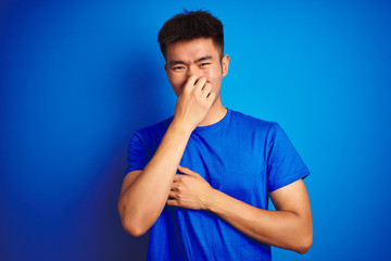 Young asian chinese man wearing t-shirt standing over isolated blue background smelling something stinky and disgusting, intolerable smell, holding breath with fingers on nose. Bad smells concept.