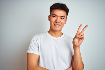 Young asian chinese man wearing t-shirt standing over isolated white background smiling with happy face winking at the camera doing victory sign with fingers. Number two.