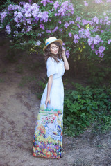 Fototapeta premium Beautiful girl in a straw hat in a lilac garden. Girl with lilac flowers in spring time. Gardening. The concept of femininity, tenderness. Bright purple flower girl. Summer flowers, Spring blossom.