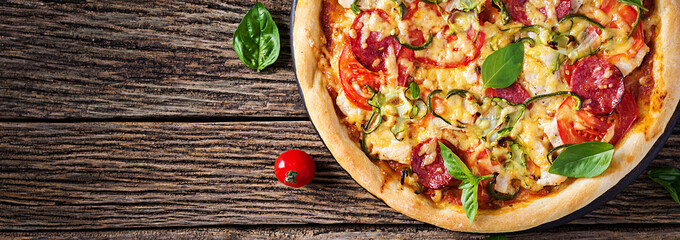 Pizza with chicken, salami, zucchini, tomatoes and herbs on vintage wooden background. Top view. Banner. Italian cuisine