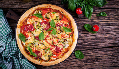 Italian pizza with chicken, salami, zucchini, tomatoes and herbs on vintage wooden background. Top view. Italian cuisine