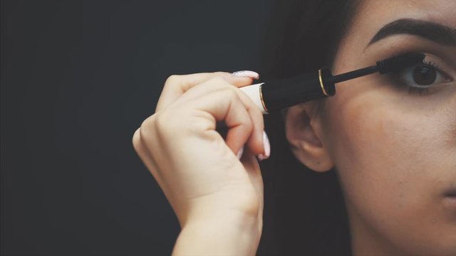 Woman applying black mascara on eyelashes with makeup brush. Young beautiful woman applying mascara makeup on eyes by brush. Portrait of brunette beauty girl applying makeup over black background.