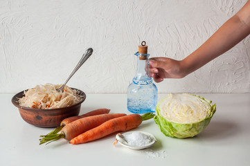 Cooking sauerkraut, a clay bowl with sauerkraut on a white table. The person’s hand holds a bottle.