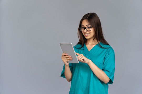 Smiling Young Indian Woman Professional Nurse Female Doctor Wear Green Uniform Glasses Holding Using Digital Tablet Isolated On Grey Studio Background, Healthcare Technology Concept, Copy Space