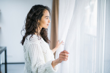 Young woman opening window curtains at home.