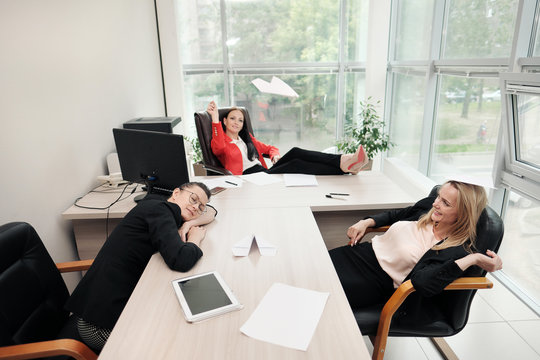 Three beautiful young girls in business suits are sitting at the office desk. Tired of work. Throw paper airplanes, and laugh. Break and rest.