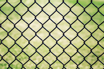 Naklejka premium Closeup of black metal netting wire mesh fence against green field meadow. Texture pattern surface background of chain link wire-mesh rabitz.