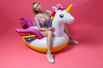 A beautiful blonde girl in a sexy sundress with slim legs in white sneakers sits on an inflatable multi-colored unicorn straightens hair on a pink isolated background. Summer vacation at the beach