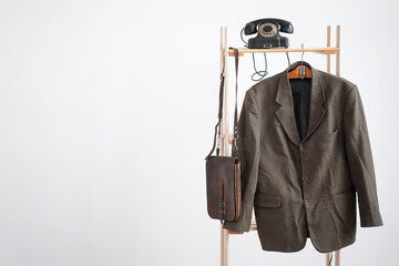 Old suit is hanging on a wooden rack with a rotary phone and old leather bag on a white wall...