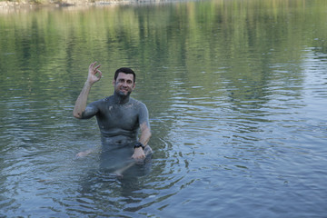 Fototapeta na wymiar A young handsome man without a shirt, his body smeared with curative mud, sits and relaxes in the water of a mountain river. Healthy lifestyle, natural procedures. The concept of unity with nature.