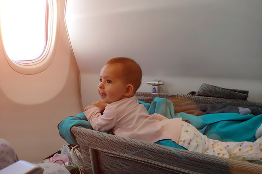 Happy Infant Baby Lyes In Special Bassinet In Airplane At His Stomach. First Flight Of The Baby, She Is Impressed And Smiles. Traveling With Child