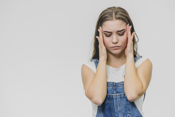 A strained woman holds her fingers in temples, feels painful emotions, suffers from headache after hard work, wears a white T-shirt, overalls, poses indoors.