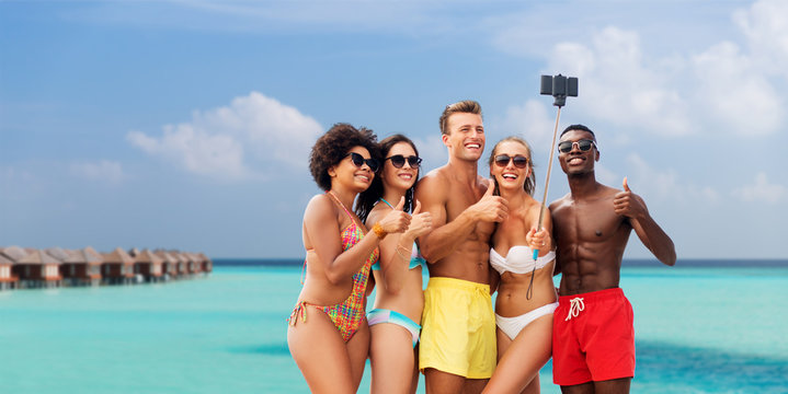 friendship, summer holidays and vacation concept - group of happy friends taking picture by smartphone on selfie stick and showing thumbs up over tropical beach background in french polynesia