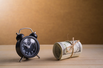 A retro clock and a roll of American currency (USD, American dollars) with 100 dollars bank notes on the outside as a symbol of plenty of money on the wooden background. 