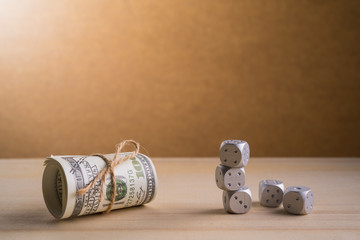 A dice and a roll of American currency (USD, American dollars) with 100 dollars bank notes on the outside as a symbol of plenty of money on the wooden background. 