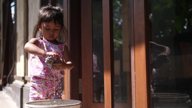 Little Indonesian child doing household chores. Tiny girl in Bali helps around the house by washing windows using a rag and bucket of water. Asian work culture in children.