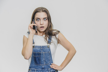 A young beautiful girl dressed in a blue denim suit and a white T-shirt. She spoke on the phone with a hand on the waist and shows a very surprised facial expression.