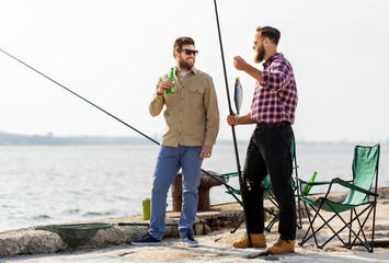 leisure and people concept - happy male friends with fishing rods, fish and beer talking on pier at sea