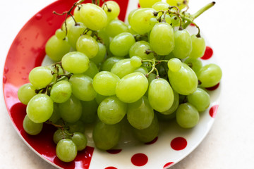 Ripe green grapes in plate on white wooden table, top view. Green grapes in a plate on the table. Green grapes in a red plate on the table.