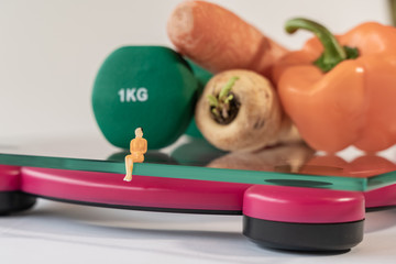 Miniature woman figure siting on the digital electronic bathroom scale for weight of human body. Fresh vegetables and green dumbbells at shallow depth of field background. Slimming concept