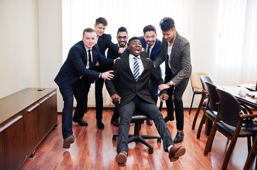 Six multiracial business mans standing at office and roll man on chair. Diverse group of male employees in formal wear having fun.