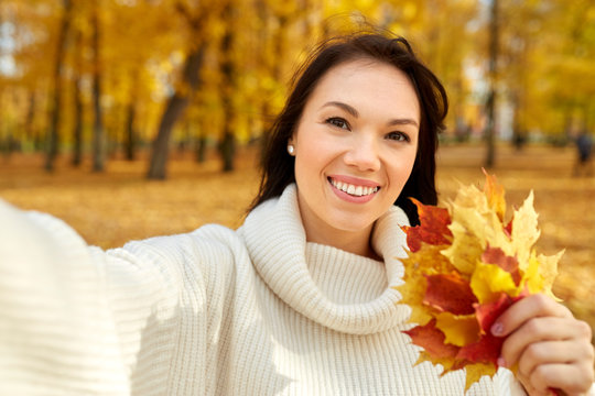 season and people concept - happy young woman with maple leaves taking selfie in autumn park