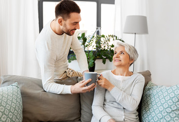 family, generation and people concept - happy smiling adult son bringing coffee to senior mother at...