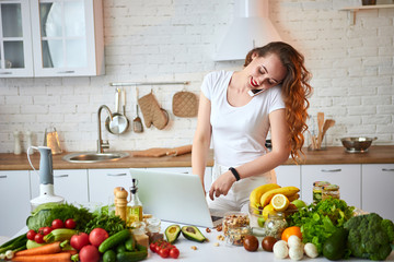 Young beautiful woman talking on smartphone and using a notebook while cooking in the modern kitchen. Healthy food and Dieting concept. Losing Weight