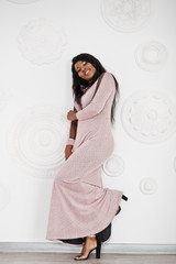 Fashionable african american model woman in pink brilliant evening dress posed against white decorative wall.