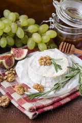 fresh round cheese on linen napkin in white red cage, branch of green grapes in bowl of natural wood, slice of figs, rosemary