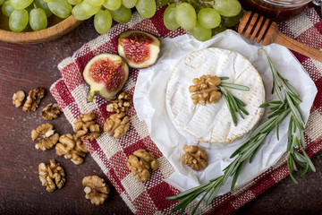 fresh round cheese on linen napkin in white red cage, branch of green grapes in bowl of natural wood, slice of figs, rosemary, top view