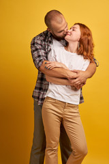 Horizontal shot of happy girlfriend and boyfriend embrace with love, pose against yellow studio wall. Bearded handsome man in checkered shirt hugs wife, stands behind. People and relationships