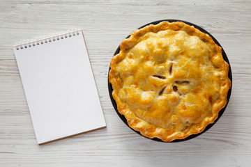 Homemade apple pie and blank notebook on a white wooden surface, top view. Flat lay, overhead, from...