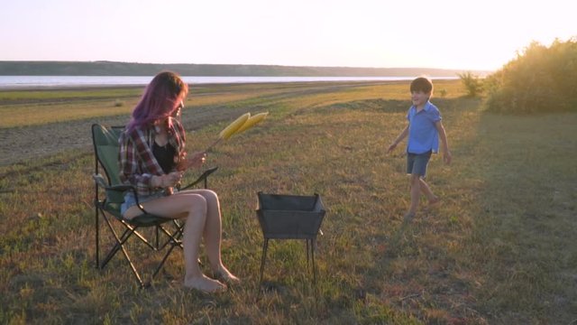 mom and son cook corn on a grill, on a picnic, on the river bank, at sunset, roasting corn on coals, 100fps