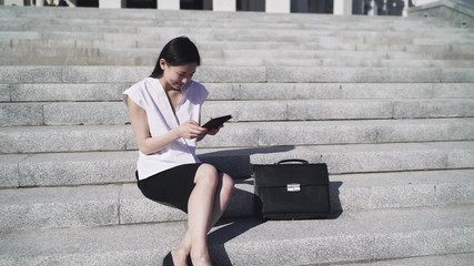 Asian business woman is sitting on the steps in city in sunny day and uses tablet pc