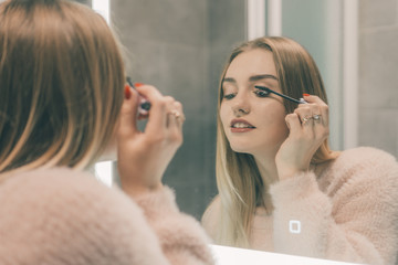 The girl in the bathroom in front of the mirror paints the eyelashes.