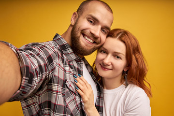 Close up portrait of young, caucasian, attractive, lovely, positive couple in shirts making selfie on mobile phone over yellow background