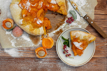 Homemade fruit pie (galette) made with fresh organic apricotes with powdered sugar on wooden table, top view. Plate with a cut piece of cake. Open pie, apricot tart. Summer pie on parchment paper.