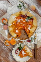 Homemade fruit pie (galette) made with fresh organic apricotes with powdered sugar on wooden table, top view. Plate with a cut piece of cake. Open pie, apricot tart. Summer pie on parchment paper.