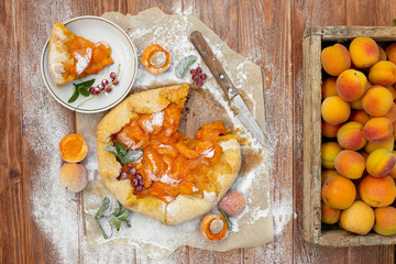 Homemade fruit pie (galette) made with fresh organic apricotes with powdered sugar on wooden table, top view. Plate with a cut piece of cake. Open pie, apricot tart on parchment paper.