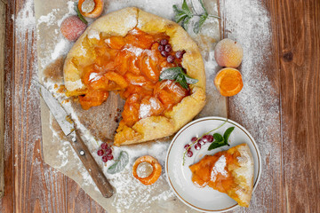 Homemade fruit pie (galette) made with fresh organic apricotes with powdered sugar on wooden table, top view. Plate with a cut piece of cake. Open pie, apricot tart on parchment paper.