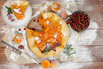 Homemade fruit pie (galette) made with fresh organic apricotes with powdered sugar on wooden table, top view. Plate with a cut piece of cake. Open pie, apricot tart on parchment paper.
