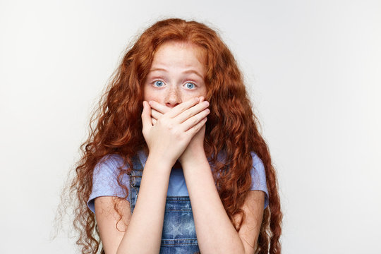 Portrait of scared cute freckles little girl with ginger hair, hears heard a terrible storry, covered mouth with palms, stands over white wall with wide open eyes with afraided expression.